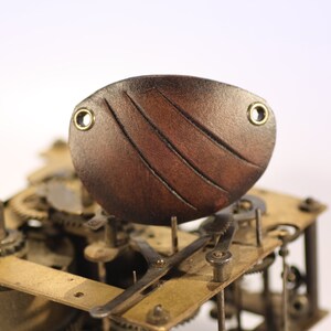 May include: A brown leather eye patch with a curved design and two brass eyelets. The eye patch is set against a backdrop of clock gears and mechanisms, suggesting a steampunk or vintage aesthetic.