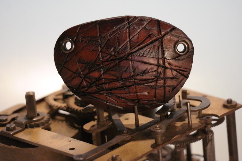 May include: A brown leather eye patch with a textured surface and two metal eyelets. The eye patch has a geometric pattern of etched lines. The background shows the gears and mechanisms of a clock.