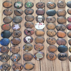 Może przedstawiać: Kolekcja 60 skórzanych naszywek z różnymi wzorami, w tym logo, symbolami i postaciami. Naszywki mają owalny kształt i brązowe skórzane tło. Niektóre wzory są czarno-białe, a inne kolorowe.