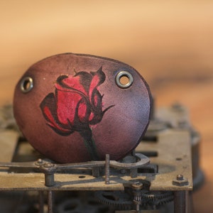 May include: A brown leather eye patch featuring a hand-painted red rose. The eye patch has two metal eyelets and is set against a blurred background of a clock mechanism.