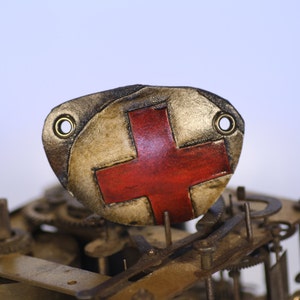 Könnte beinhalten: Nahaufnahme einer roten Kreuz auf einer goldfarbenen Metallplakette mit zwei Löchern. Die Plakette ist an einem rostigen Metallgetriebe befestigt.