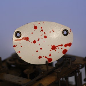 May include: White, oval-shaped metal piece with two eyelets and red splatter paint. The piece is positioned above a complex mechanical structure, possibly clock gears. The background has a gradient of blue and purple.