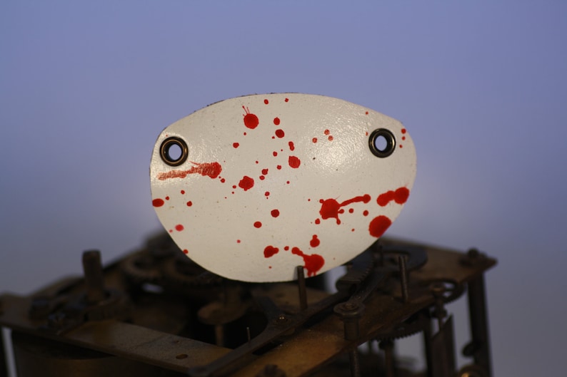 May include: White, oval-shaped metal piece with two eyelets and red splatter paint. The piece is positioned above a complex mechanical structure, possibly clock gears. The background has a gradient of blue and purple.