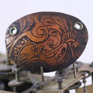 Puede incluir: Un parche para el ojo de cuero marrón con un diseño floral grabado. El parche para el ojo tiene dos ojales metálicos y está colocado sobre un fondo de engranajes de reloj. El cuero es de color marrón cálido y el diseño grabado es marrón oscuro.