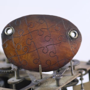 Könnte beinhalten: Ein braunes Lederpuzzleteil mit dunklen Umrissen von Puzzleteilen. Das Lederteil hat zwei Metallösen und ist vor dem Hintergrund von Uhrwerkzahnrädern und Mechanismen angeordnet. Das Teil ist oval geformt.