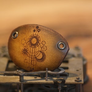 Könnte beinhalten: Eine braune Leder-Augenklappe mit einem Himmelsdesign, einschließlich Sonnen, Monde und Sterne. Die Augenklappe hat zwei Metallösen. Das Design ist in das Leder geätzt.