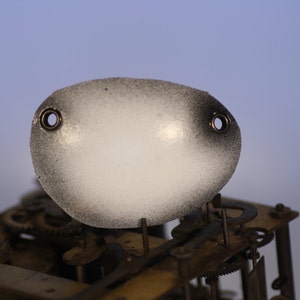May include: Oval-shaped clock part with a frosted white center and black speckled edges. Two small holes are visible near the top. The part is positioned above the intricate gears and mechanisms of a clock.