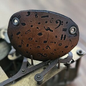 Op de afbeelding: Een bruine leren sleutelhanger met gegraveerde muzieknoten. De sleutelhanger is bevestigd aan een metalen mechanisme.