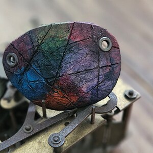 Pode incluir: Um objeto oval colorido com uma superfície rachada e texturizada, apresentando um mosaico de tons de arco-íris. Dois ilhós de metal são visíveis. O objeto repousa sobre um conjunto mecânico complexo de cor latão, sugerindo um relógio ou dispositivo semelhante.