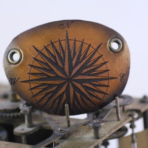 Pode incluir: Uma tapa-olho de couro castanho com um desenho de rosa dos ventos em preto. A tapa-olho tem dois ilhós de metal e está sobre um fundo de engrenagens de relógio. A rosa dos ventos tem marcações direcionais.