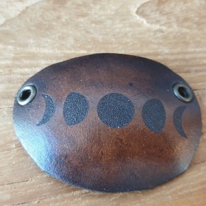 May include: A brown leather bracelet with a moon phase design and two silver metal rivets.