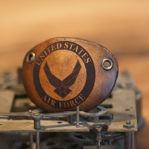 Peut inclure: Un patch en cuir marron avec l'inscription "UNITED STATES AIR FORCE" encerclant un emblème noir d'oiseau. Le patch comporte des œillets métalliques et se détache sur un fond flou de mécanisme d'horloge.