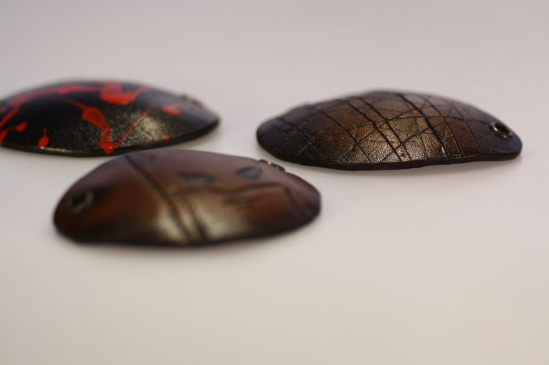 May include: Three oval-shaped leather pieces in varying shades of brown and black. One is black with red paint splatters, another is a dark brown with a grid pattern, and the third is a lighter brown with etched lines. Each has a small metal ring.