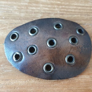 May include: Oval-shaped, brown leather eye patch with multiple metal grommets. The eye patch is designed with a distressed finish, and the grommets are evenly spaced across the surface. The background is a light brown wooden surface.