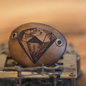 Pode incluir: Uma tapa-olho de couro castanho com um desenho de diamante. A tapa-olho tem dois ilhós de metal e é colocada sobre um fundo desfocado de um mecanismo de relógio e uma superfície de madeira.