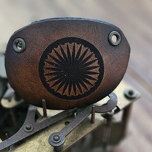 Op de afbeelding: Een bruine leren sleutelhanger met een zwart cirkelvormig ontwerp in het midden. Het ontwerp heeft een zonnestralenpatroon.