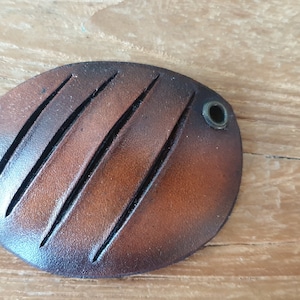 Könnte beinhalten: Ein braunes Leder-Fisch-Patch mit zwei Metallösen. Das Patch hat eine strukturierte Oberfläche mit fünf parallelen Linien, die in das Leder geschnitten sind.