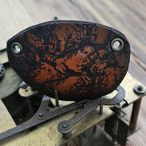 Pode incluir: Uma tapa-olho de couro castanho com um design marmorizado preto e duas ilhós de metal. A tapa-olho está colocada sobre um fundo de engrenagens de relógio e tábuas de madeira, mostrando uma estética steampunk.