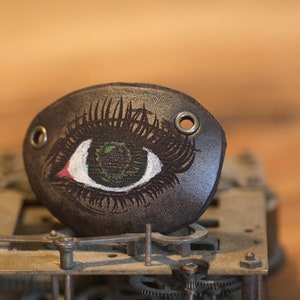 May include: A dark brown leather eye patch with a detailed eye design. The eye features a green iris, white highlights, and a red accent. The patch has two metal grommets and is set against a blurred background.