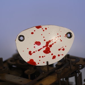 May include: A white metal clock face with red paint splatters. The clock face is oval shaped and has two small holes on either side.