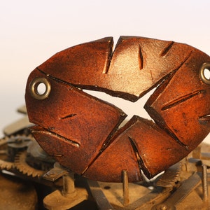 Könnte beinhalten: Ein braunes, ovales Keramikstück mit zwei Messingösen. Das Stück ist auf einem Metallsockel mit Zahnrädern montiert.