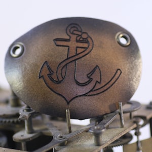 May include: A brown leather eye patch with a laser-etched anchor design. The eye patch has two metal grommets and is set against a blurred background of clock gears. Nautical themed accessory.