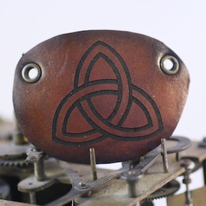May include: A brown leather patch with a black Celtic knot design. The patch has two metal eyelets and is set against a background of clock gears. The leather has a textured appearance.