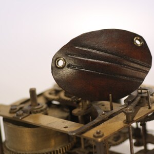 May include: Close-up of a vintage clock mechanism with a brown leather cover. The cover has three decorative lines and two brass eyelets. The mechanism is made of brass and has several gears and levers.