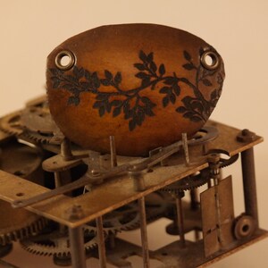 Peut inclure: Une pièce de cuir ovale marron avec un motif floral noir, probablement un composant d'horloge, est posée sur un ensemble complexe d'engrenages et de pièces métalliques. La pièce a deux œillets et une esthétique vintage détaillée.