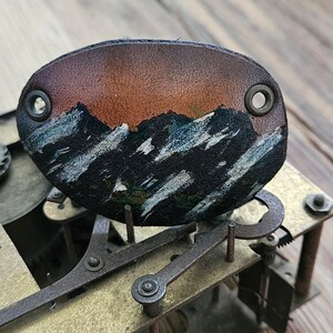 Op de afbeelding: Een leren patch met een geschilderd berglandschap. De bergen zijn geschilderd in tinten blauw, wit en zwart, met een zonsondergangshemel op de achtergrond. De patch heeft twee metalen oogjes boven en onder.