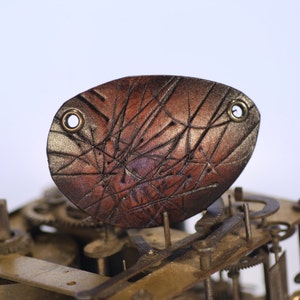 May include: A brown leather patch with a distressed, textured surface and two brass eyelets. The patch is attached to a metal mechanism with gears.