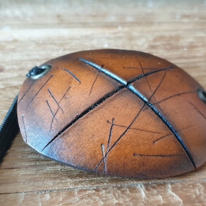 May include: A brown leather eye patch with a black elastic strap. The eye patch is oval-shaped with etched lines and metal grommets. The leather has a distressed appearance.