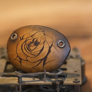 May include: A brown leather cuff with a rose design. The cuff has two metal eyelets.