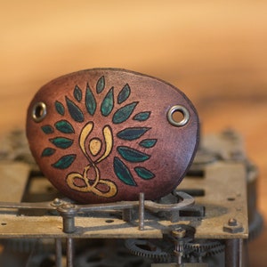 May include: Brown leather eye patch featuring a painted tree of life design in green and gold. The patch has two metal eyelets for a strap. The background is a vintage clock mechanism.