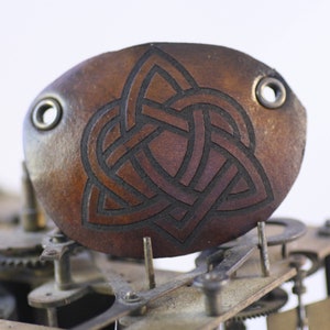 Pode incluir: Uma pulseira de couro castanho com um desenho de nó celta. A pulseira tem dois ilhós de metal e está colocada sobre um fundo de engrenagens de relógio. O nó é de cor castanho escuro.