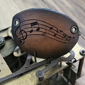 Op de afbeelding: Een bruine leren sleutelhanger met een muzieknootontwerp. De sleutelhanger is bevestigd aan een metalen tandwielmechanisme.