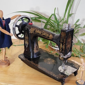 May include: A black vintage sewing machine with gold floral decals and the text "ROTARY SPECIAL" printed on the side. The machine features a large silver handwheel and is accompanied by two wooden articulated artist mannequins on a wooden surface.