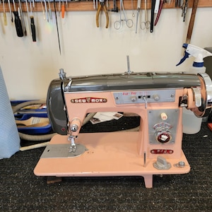 Pode incluir: Uma máquina de costura vintage rosa e cinza Sew Mor Zig-Zag. A máquina tem uma roda e um painel de controle prateados. A máquina está sobre uma superfície preta. Ferramentas penduradas na parede ao fundo.