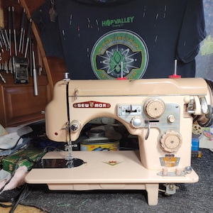 May include: A vintage beige Sew Mor Deluxe sewing machine with chrome accents. The machine features multiple dials, a needle, and a flat base. The Sew Mor logo is visible on the side. The background includes tools and a t-shirt.