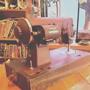 Puede incluir: Una máquina de coser marrón vintage con una rueda grande y una base rectangular. La máquina tiene un cuerpo de metal y una aguja, con la marca "New Home" visible en el lateral. La máquina está sobre una superficie de madera.
