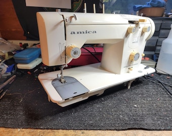 May include: Off-white Amica sewing machine with yellow knobs and the text "amica" on the front. The machine has a needle, a presser foot, and a flat base. The model name "Super 5000" is visible.