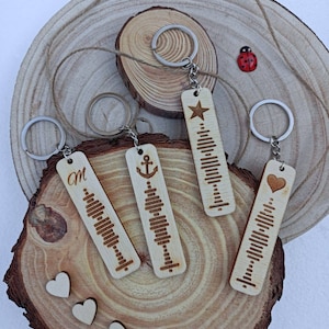 Könnte beinhalten: Vier Holzschlüsselanhänger mit verschiedenen Designs. Jeder Schlüsselanhänger zeigt ein Schallwellenmuster und ein einzigartiges Symbol: einen Stern, einen Anker, den Buchstaben "M" und ein Herz. Die Schlüsselanhänger werden auf einer Holzoberfläche präsentiert.