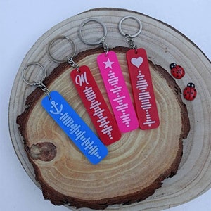 Pode incluir: Quatro chaveiros diferentes com designs personalizados. Os chaveiros são feitos de plástico vermelho, rosa e azul. Cada chaveiro tem um design branco. Os designs incluem uma estrela, um coração, a letra M e uma âncora.