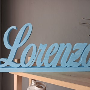 Könnte beinhalten: Hellblaues Holzschild mit dem Namen "Lorenzo" in Schreibschrift. Das Schild ist freistehend und steht auf einem hellen Holzregal. Der Hintergrund ist eine neutrale graue Wand.