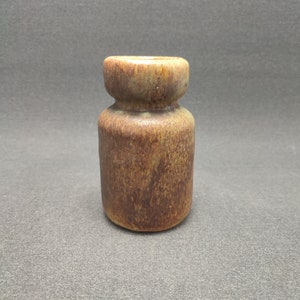 May include: A small, cylindrical ceramic vase with a rounded top and a wider base. The vase has a mottled brown and tan glaze, giving it a rustic appearance. The vase is approximately 10 cm tall.