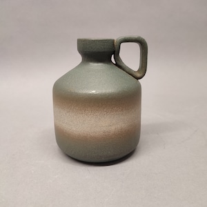 Puede incluir: Un pequeño jarrón de cerámica verde con un diseño de rayas blancas y marrones. El jarrón tiene un asa y un cuerpo redondeado.