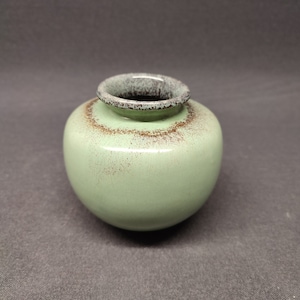 Może przedstawiać: Mały, okrągły, zielony ceramiczny wazon z brązową krawędzią. Wazon ma pstrokatą glazurę.