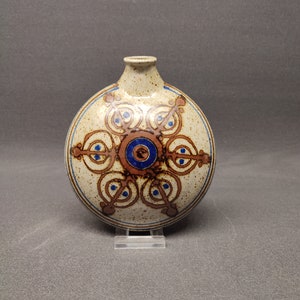 Puede incluir: Un jarrón redondo de cerámica con un diseño geométrico marrón, blanco y azul. El jarrón tiene un esmalte moteado y una pequeña abertura en la parte superior.