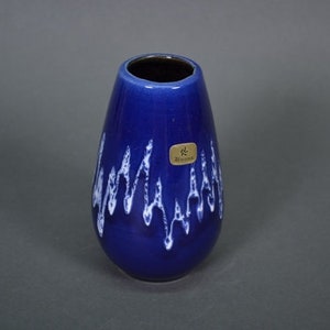 May include: A blue ceramic vase with a white glaze design. The vase has a rounded bottom and a narrow opening. It has a label that says "* Keramik"