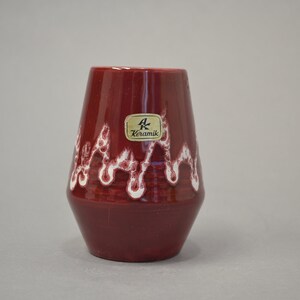 May include: A red ceramic vase with white glaze, featuring a wavy design. The vase has a label that reads "AK Keramik".